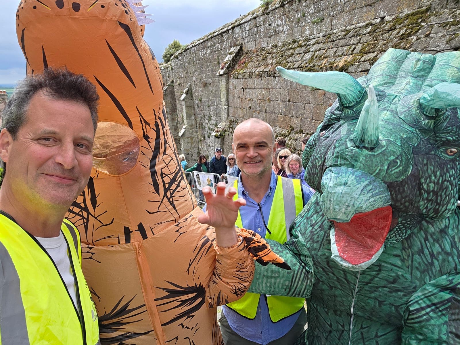 Jim in a hi-vis marshalling the Gold Hill Cheese Race in Shaftesbury, beside two people in inflatable dinosaur costumes