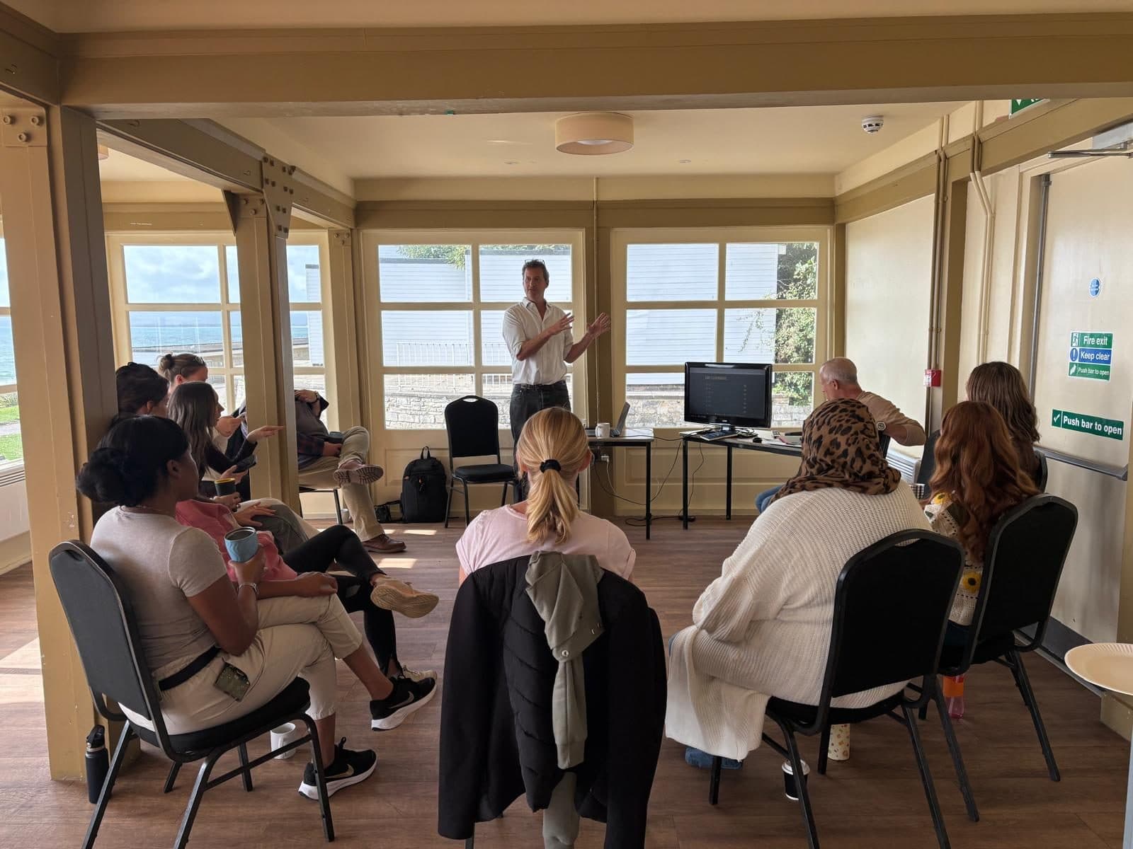 Jim Chetwode presenting to a seated group of eleven communications staff in a seafront room with the sea visible through the windows